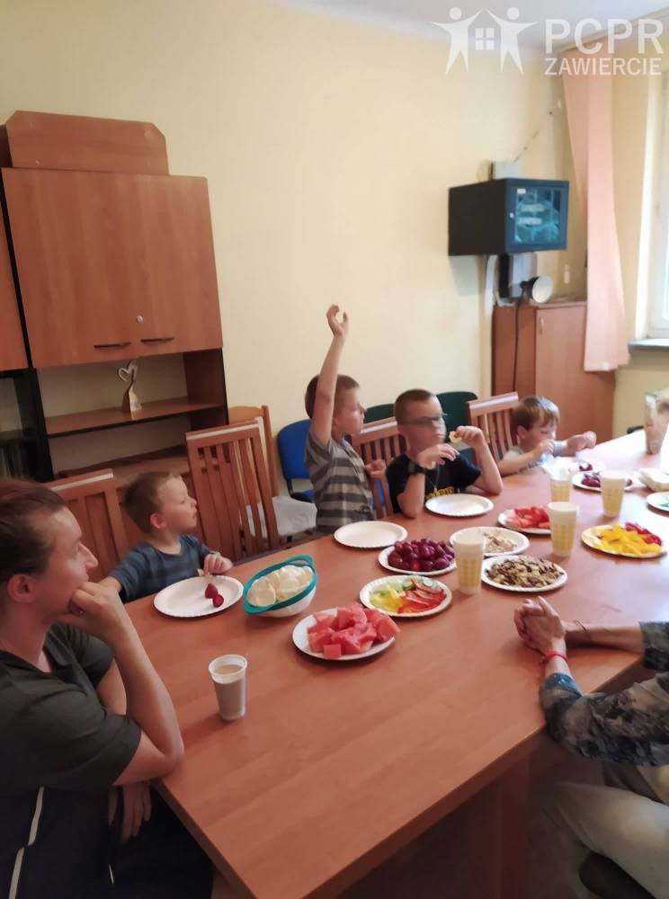 Zdjęcie: Grupa dzieci siedzi przy stole, na którym w pojemnikach poustawiane są pokrojone produkty spożywcze, a kobieta tłumaczy, a chłopiec zgłasza się do odpowiedzi